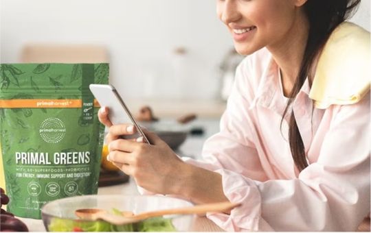 Primal greens on a kitchen counter next to a woman on her phone.